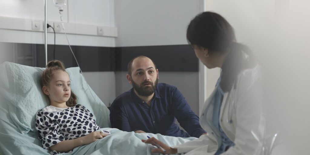 Mädchen im Krankenhausbett, daneben Vater und Ärztin im Gespräch
