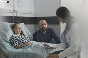 Mädchen im Krankenhausbett, daneben Vater und Ärztin im Gespräch